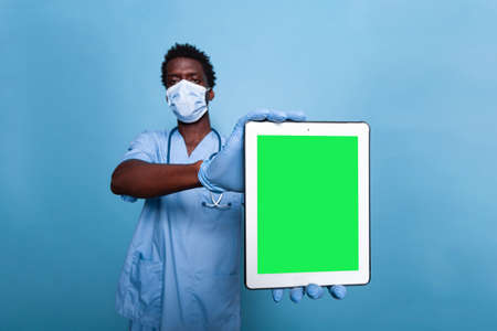 Medical nurse holding vertical green screen on tablet in studio. Healthcare assistant with covid 19 face mask and stethoscope having gadget with chroma key, showing isolated mockup template.の写真素材