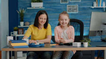 Portrait of mother and daughter sitting at desk for homework and online classes. Young child preparing to do tasks for remote lessons and courses with parent while looking at camera.の写真素材