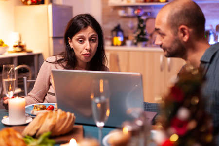 Joyful couple browsing on social media reading entertainment message while sitting at dining table in xmas decorated kitchen. Happy family celebrating christmas holiday enjoying winter season togetherの写真素材