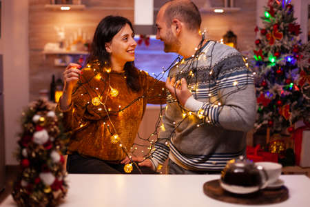 Amused couple having fun with christmas tree light decorating kitchen enjoying spending wintertime together. Happy family celebrating santa-claus festive season. Christmastime traditionの写真素材