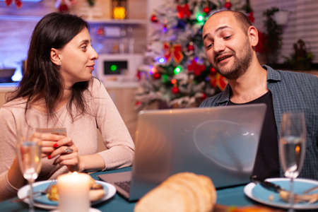 Cheerful couple shopping online xmas gift present paying with credit card on laptop computer sitting at christmas table in x-mas decorated kitchen. Happy family celebrating winter holidayの写真素材