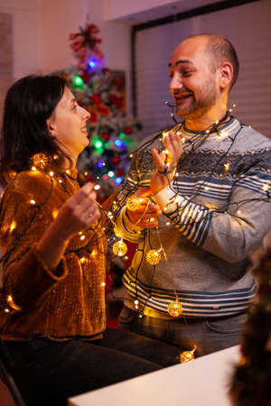 Amused couple having fun with christmas tree light decorating kitchen enjoying spending wintertime together. Happy family celebrating santa-claus festive season. Christmastime traditionの写真素材