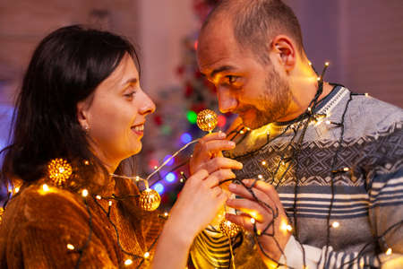 Happy amused married couple stucking in christmas tree lights enjoying spending winter holiday together. Cheerful family decorating kitchen celebrating christmastime. Santa-claus traditionの写真素材