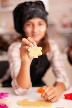 Portrait of grandchild wearing apron looking into camera while holding cookie dough using baking shape preparing homemade dessert. Child celebrating christmas holiday in xmas decorated kitchenの写真素材