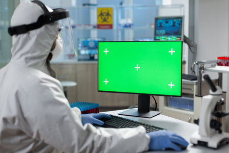 Biologist man with protection ppe medical suit looking at mock up green screen chroma key computer with isolated display while working in biochemistry hospital laboratory. Covid19 vaccine developmentの写真素材