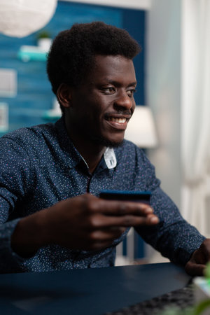 Teenager consumer making online shopping on retail store paying order with credit card. Man sitting at desk in living room doing online payment with electronic money using computerの写真素材