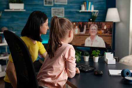 Granddaughter sitting beside parent talking with retired grandmother during online video call meeting conference enjoying spending time together. Teleconference call on computer screenの写真素材