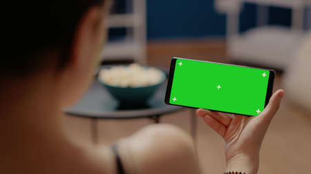Close up of mobile phone with horizontal green screen on display. Young woman sitting at home...