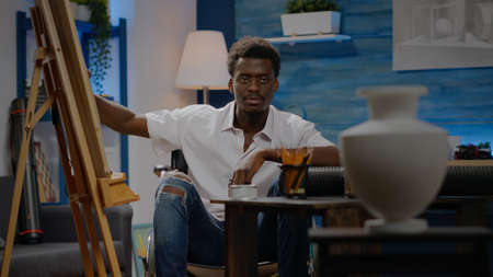 African american artist designing art drawing of white vase in workshop space. Black young person sitting in wheelchair working on modern masterpiece for successful galleryの写真素材