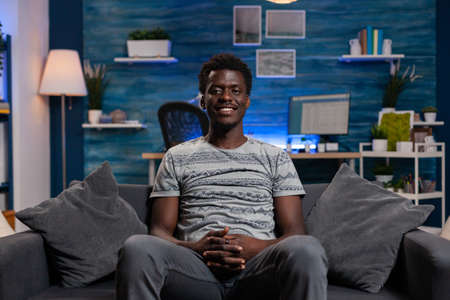 Portrait of african american young employee sitting on sofa looking into camera while working remote...