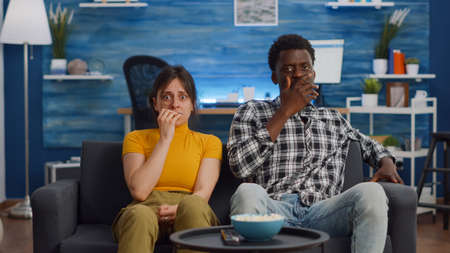 POV of shocked interracial couple watching scary movie on television at home. Multi ethnic husband and wife being afraid and covering eyes while looking at camera in living roomの写真素材