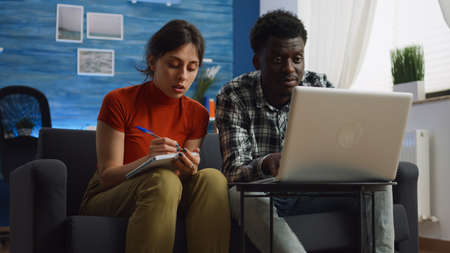 Young interracial couple calculating taxes and paperwork using laptop and notebook in living room....