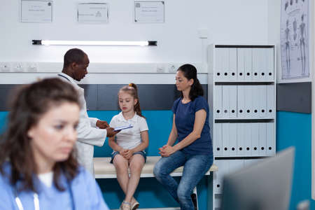 Practitioner woman asisstant typing disease symptoms on computer working at healthcare treatment during clinical appointment in hospital office. In background doctor examining sick girl patientの写真素材