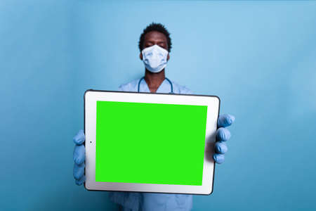 Healthcare assistant showing tablet with horizontal green screen. Medical nurse with face mask,...