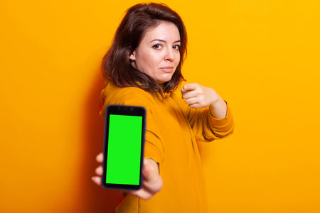 Woman showing mobile phone with vertical green screen at camera in studio. Adult pointing at mobile phone with isolated mockup template on display. Person standing over orange backgroundの写真素材