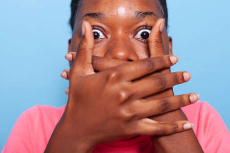 Closeup of african american young woman covering mouth having shocked facial expressions while standing in studio with blue background. Portrait of terrified teenager looking at cameraの写真素材