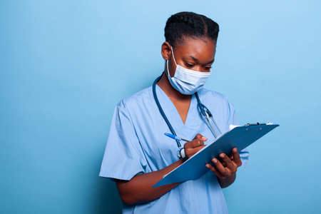 African american therapist nurse wearing protective medical face mask against virus writing healthcare treatment on clipboard in studio with blue background. Medicine conceptの写真素材