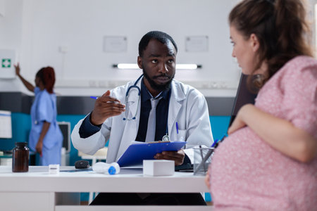Medical physician consulting woman expecting child at desk in cabinet. General practitioner talking to pregnant patient about pregnancy and healthcare, specialist giving advice.の写真素材