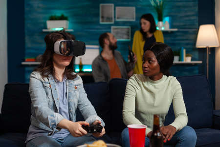 Player woman with virtual reality headset holding gaming joystick playing videogames during online competition. Group of multi-ethnic friends supporting her enjoying free time togetherの写真素材