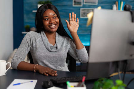 Young woman waving at video call camera on computer, talking to colleagues at home. Person attending...