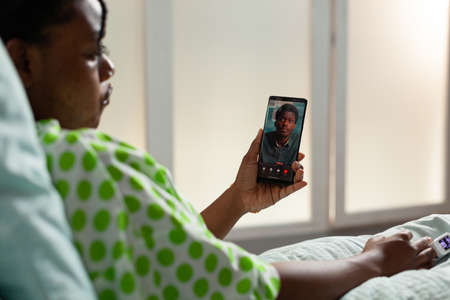 African american young patient holding smartphone explaining sickness expertise to remote friends during online videocall meeting conference. Sick woman lying in bed in hospital officeの写真素材