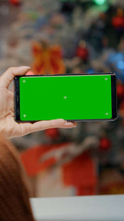 Young woman using smartphone in festive kitchen decorated with ornaments and tree. Adult holding green screen background device for digital seasonal holiday celebration dinner at homeの写真素材