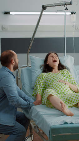 Young man panicking while pregnant wife having contractions in hospital ward. Caucasian husband getting medical assistance help for screaming wife in pain. Childbirth agony and acheの写真素材