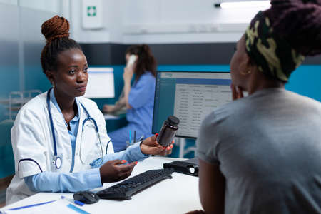 African american physician holding antibiotic bottle in hand discussing disease symptoms with patient explaining medication treatment during clinical consultation. Doctor working in hospital officeの写真素材