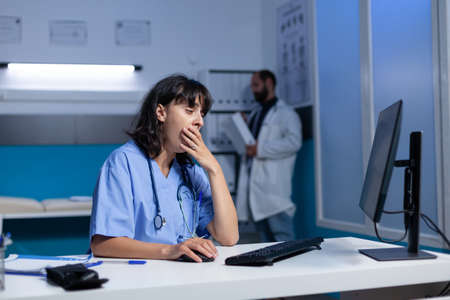 Exhausted nurse using computer on desk while falling asleep, working late. Medical assistant looking at monitor and feeling burnout after overtime work at night in healthcare office.の写真素材