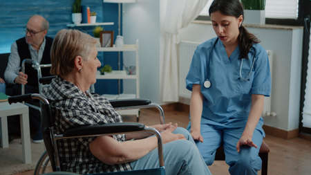 Disabled senior woman in nursing home talking to nurse for healthcare examination and checkup. Medical assistant giving advice for recovery to elder patient with disability in wheelchair.の写真素材