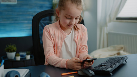 Little girl looking at smartphone display and smiling at desk. Young child in elementary school using mobile phone with touch screen for online remote class lesson. Distance educationの写真素材