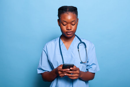 African american practitioner nurse holding smartphone browsing medical information on internet working at sickness expertise in studio with blue background. Medicine conceptの写真素材
