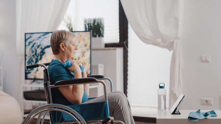 Old woman with physical disability lifting dumbbells while looking at tablet with training video. Senior adult sitting in wheelchair, doing exercise with weights and watching online lessonの写真素材