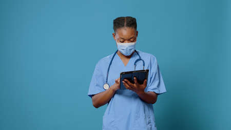 Medical assistant using digital tablet with touch screen in studio. Woman nurse working with modern device and technology while wearing face mask and uniform. Specialist with gadgetの写真素材