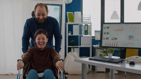 Cheerful employee taking break pushing wheelchair with his paralysed invalid disabled colleague...