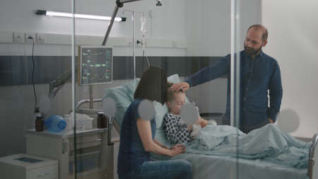 Hospitalized sick child patient falling asleep recovering after illness surgery during medical consultation in hospital ward. Worried parents discussing therapy treatment waiting for disease expertiseの写真素材