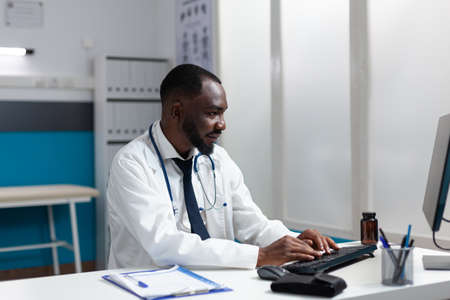 African american therapist doctor typing medical expertise on computer working at healthcare treatment in hospital office. Man physician analyzing online pharmaceutical prescriptionの写真素材