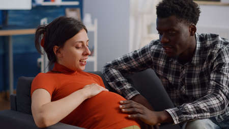 Close up of interracial couple with pregnancy at home holding hands on belly. Mixed race young...