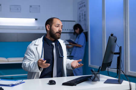 Doctor using video call on computer for remote communication, working late in office. Medic talking...