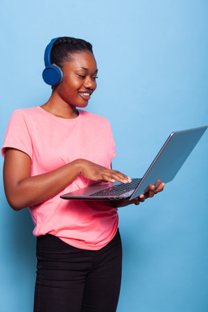 Portrait of african american student with headphones listening music searching playlist on laptop...