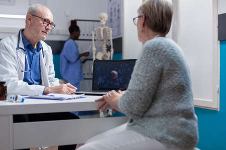 Man doctor explaining coronavirus bacteria illustration on laptop to woman in cabinet. Medical specialist and patient looking at computer screen with visual representation of covid 19 pandemic.の写真素材