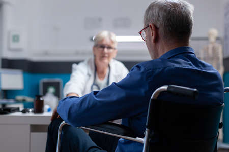 Close up of patient sitting in wheelchair at healthcare consultation with physician in cabinet. Senior man with disability meeting with medic for checkup appointment to help with illness.の写真素材