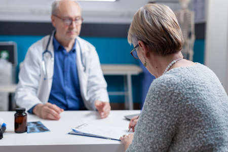 Senior woman with disease signing medical appointment papers with specialist in cabinet. Sick patient doing signature on checkup visit document to receive prescription treatment.の写真素材