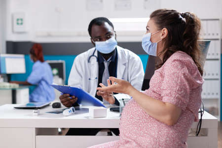 Woman expecting child and having discussion with physician about pregnancy and healthcare. General practitioner giving medical advice to pregnant patient during coronavirus pandemicの写真素材