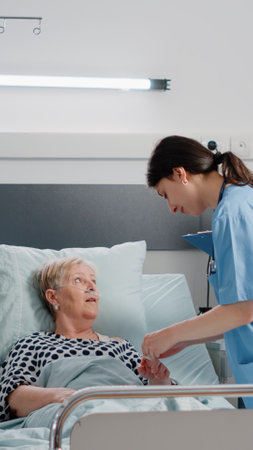 Doctor and nurse visiting patient with illness in hospital ward. Team of specialists doing consultation for elder woman with nasal oxygen tube and IV drip bag in bed. Medical recoveryの写真素材