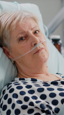 Portrait of elder woman with nasal oxygen tube laying in bed. Aged patient with disease waiting on...