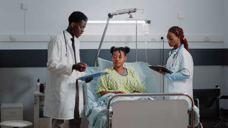 Man and woman talking to ill patient about treatment and medicine in hospital ward. Team of healthcare specialists doing consultation with young adult using tablet and documents.の写真素材