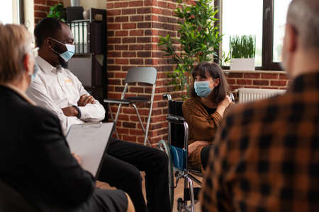 People and woman sitting in wheelchair having conversation at aa meeting. Multi ethnic patients attending conversation with psychiatrist at support group therapy during covid 19 pandemic.の写真素材