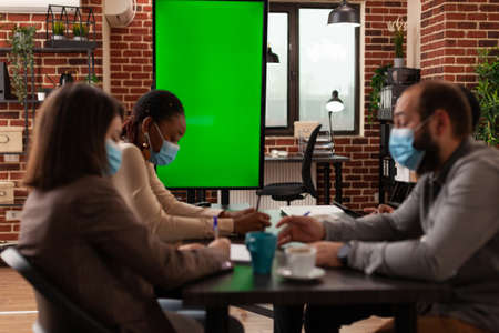 Multi-ethnic businesspeople with medical face mask against coronavirus working at business presentation in startup company meeting office. Mock up green screen chroma key monitor with isolated displayの写真素材