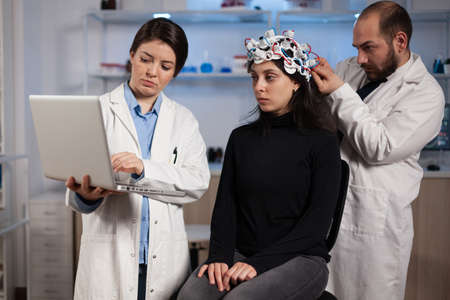 Specialist doctor holding laptop computer showing tomography to woman patient while neurologist man adjusting eeg scanner during neurology experiment. Team of researchers analyzing brain activityの写真素材
